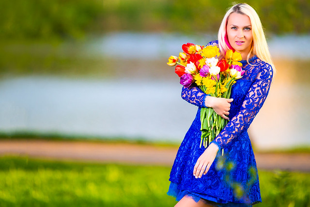 Fashion Concepts. Tranquil Positive Caucasian Girl with Bunch of Fresh Colorful Tulips. Posing in Blue Dress Outdoors During Spring Time.