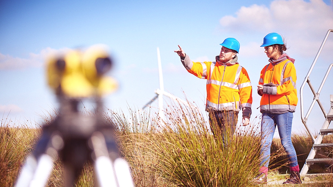 A young man points out of frame, standing next to a young woman. Both are wearing hard hats and high-vis jackets, and are standing near a wind farm.